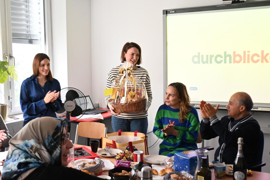 Team und Teilnehmer*innen sitzen um einen Tisch und überreichen der Kursleitung einen Geschenkkorb